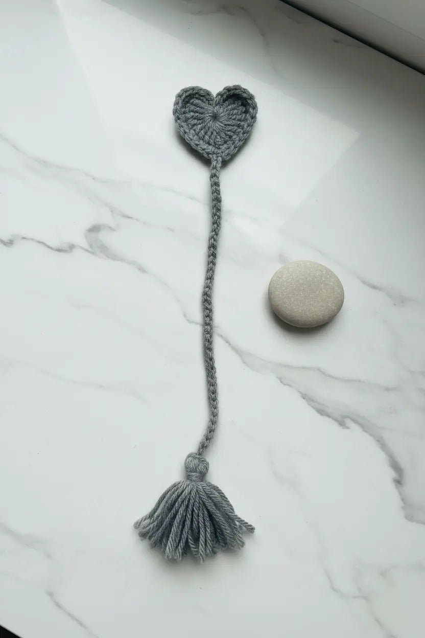 A gray crocheted bookmark with a heart-shaped top and tassel, displayed on a white marble surface next to a small round stone.