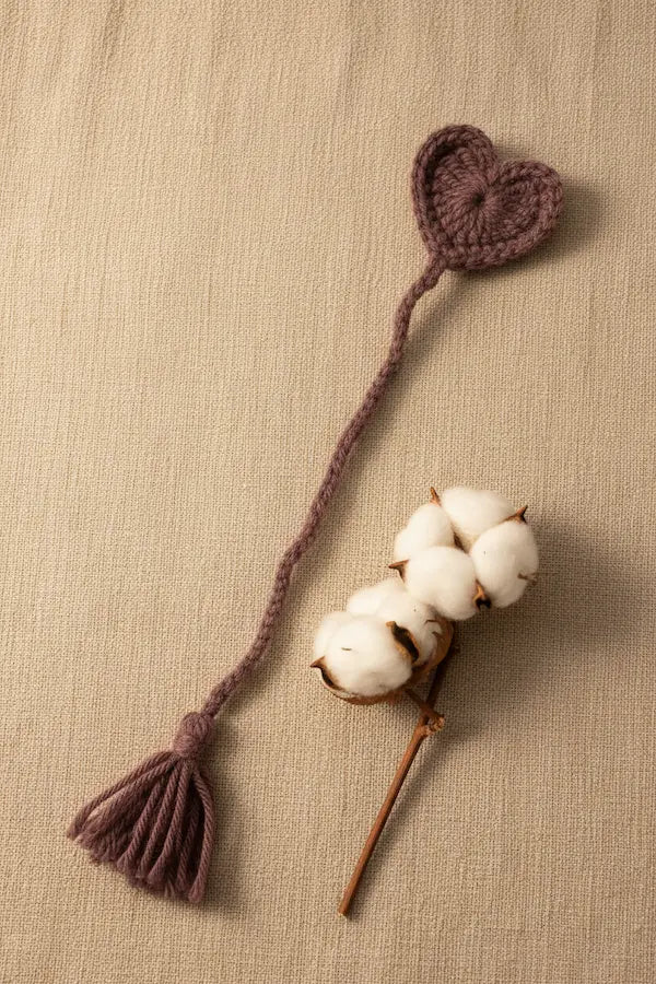 A handmade knitted heart-shaped tassel accessory in a muted mauve-brown color, styled on a textured beige linen background next to a cotton boll stem.