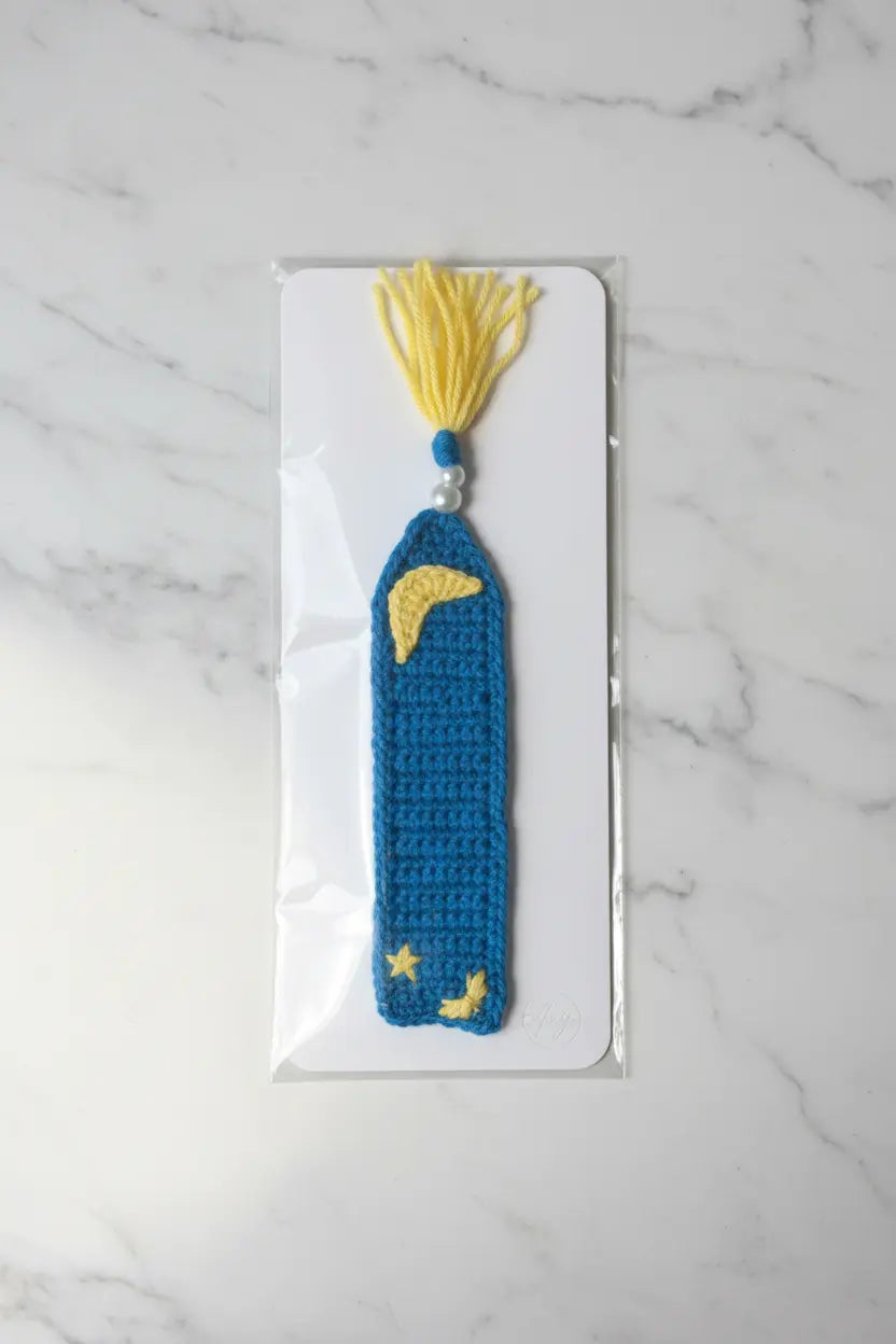 A crocheted blue book bookmark with yellow tassels and embroidered yellow moon and star details, displayed on a white card over a light gray marble background.