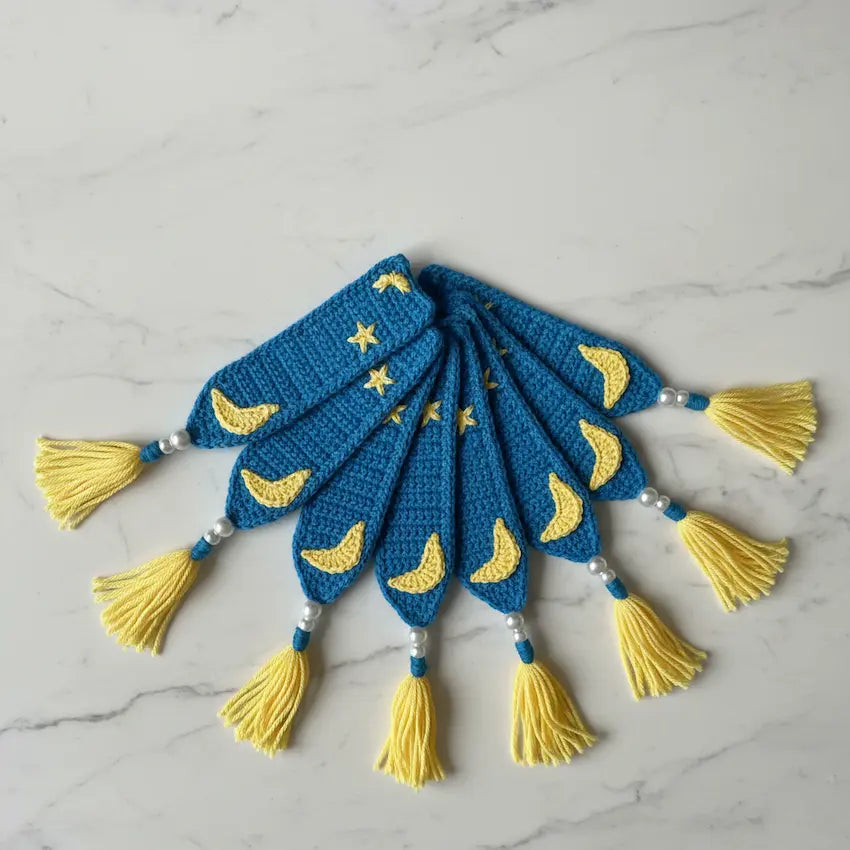 A set of five crocheted blue bookmarks with yellow crescent moon and star patterns, finished with yellow tassels and white pearl beads, arranged on a light gray marble surface.