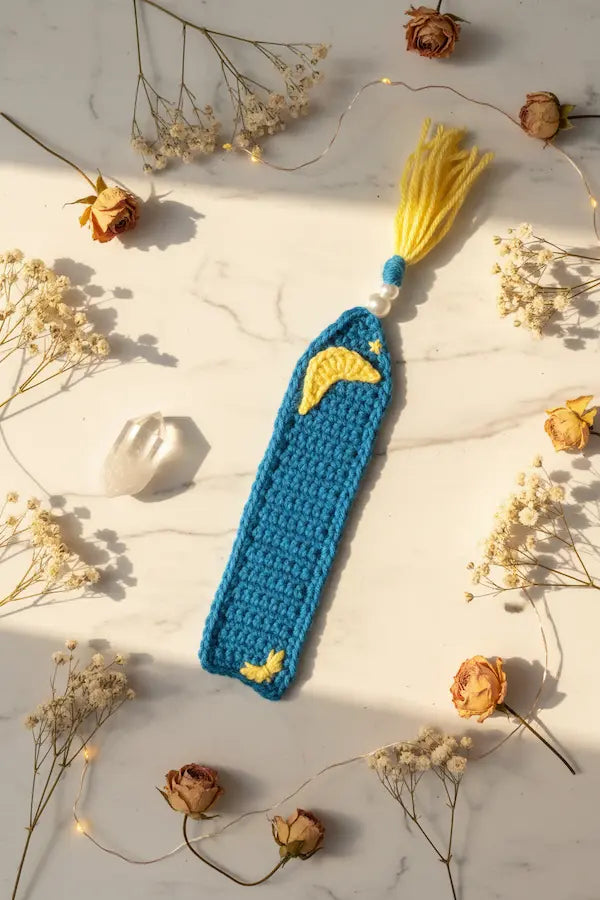 A crocheted bookmark with a blue body, yellow crescent moon and sun appliques, and a yellow tassel, styled on a marble surface with dried flowers and fairy lights.