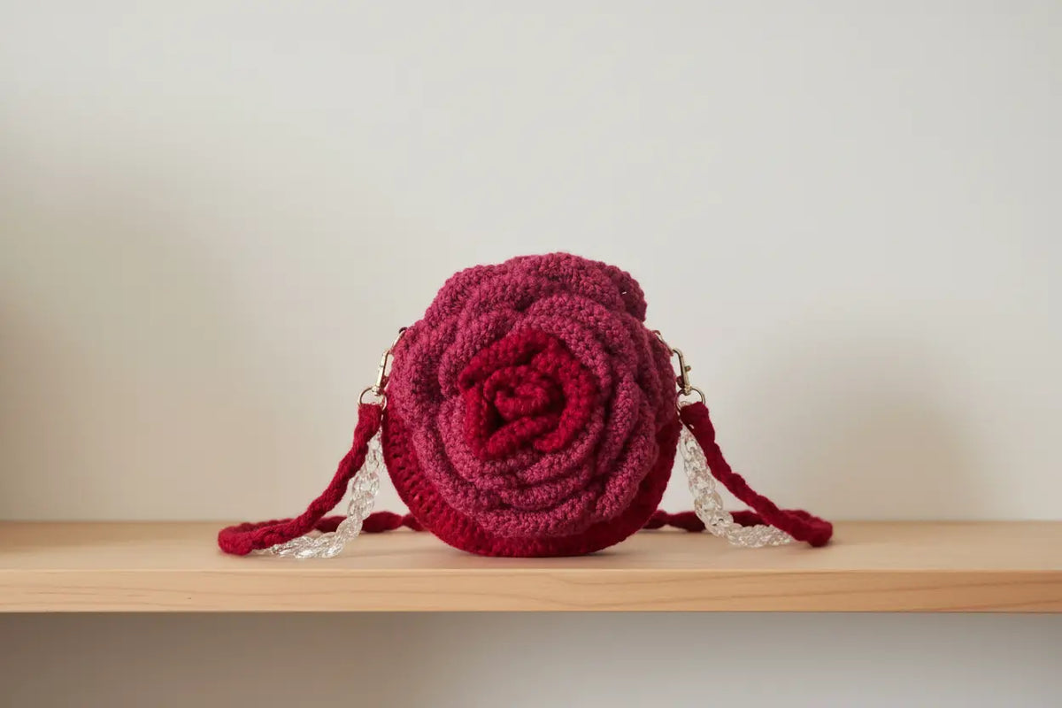 A deep burgundy crocheted rose-shaped shoulder bag with white lace side straps, resting on a light wood shelf against a plain beige wall.