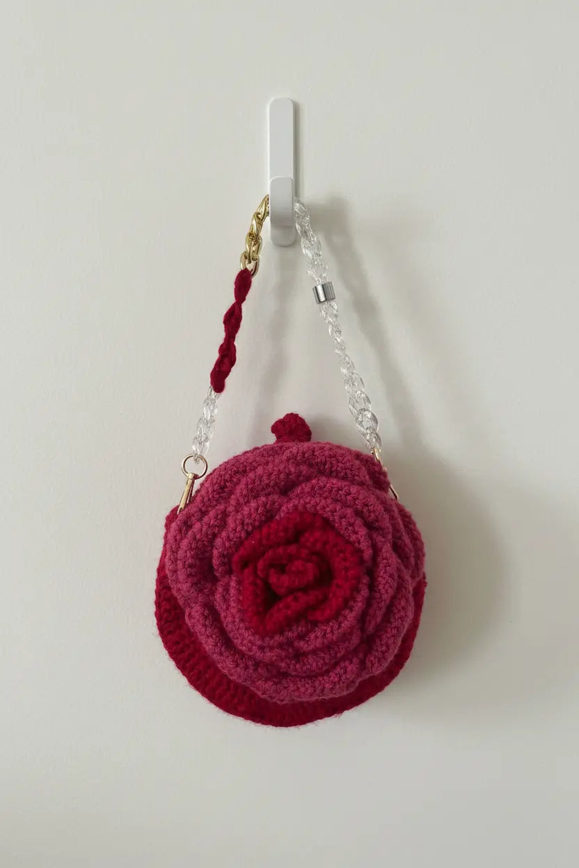 A small round crocheted rose-shaped handbag in deep magenta pink, hanging from a white wall hook. The bag features a twisted dark red handle, a clear beaded chain strap with gold-toned metal hardware, and a small top loop.