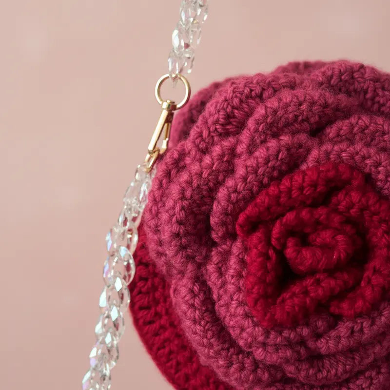 A close-up of a crocheted purse shaped like a rose in deep burgundy and magenta, featuring a clear rhinestone-embellished strap with a gold clasp against a soft pink background.