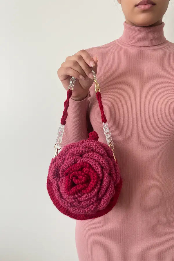 A person wearing a pink ribbed turtleneck sweater holds a round, rosette-shaped handbag made of knitted fabric in deep magenta and burgundy tones. The bag has a woven red handle with clear rhinestone-like links and gold-tone hardware, shown against a plain light gray background.