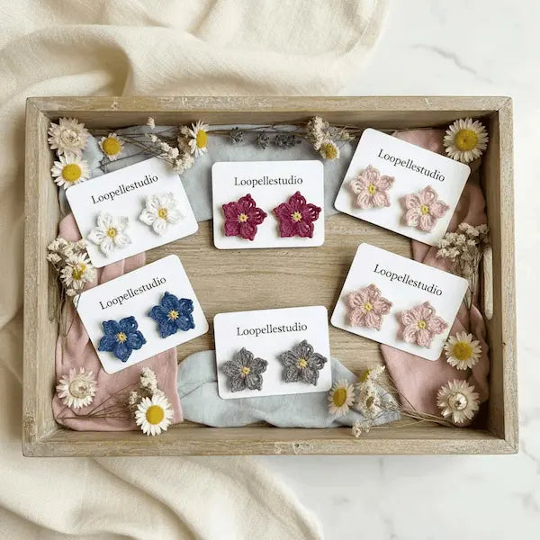 Five pairs of flower-shaped stud earrings by Loopellestudio arranged in a rustic wooden tray with pink fabric, dried flowers, and cream cloth. The earrings come in white, blue, burgundy, light pink, and gray, each with a small yellow center.