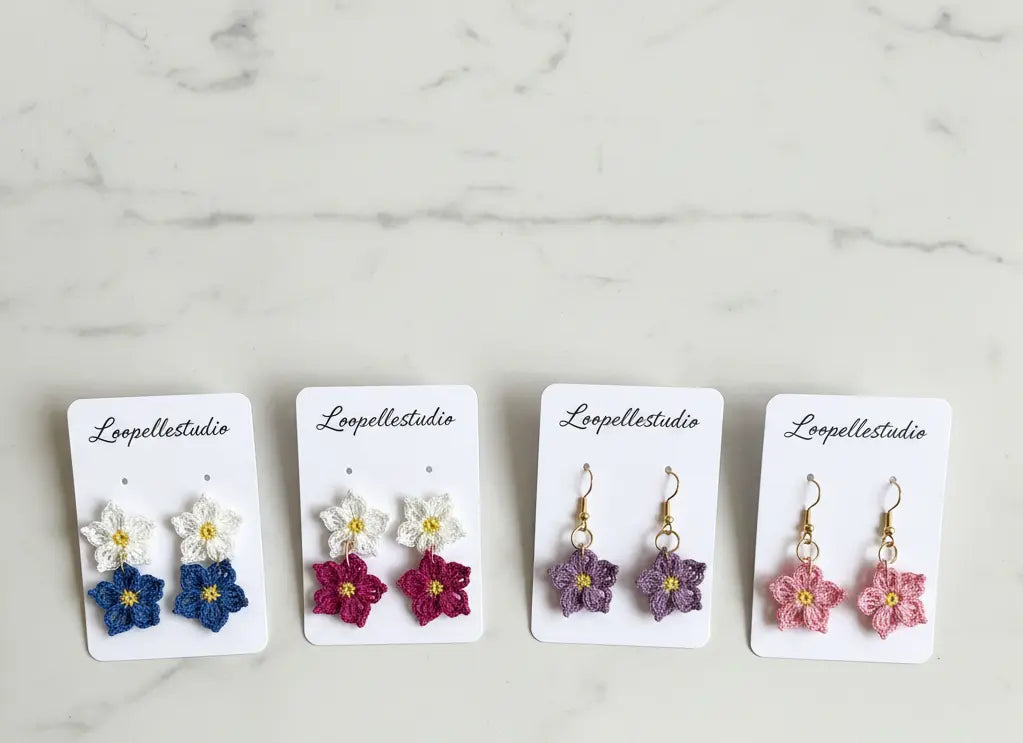 Four pairs of handmade flower dangle earrings on white display cards, arranged on a light marble surface. The pairs are blue and white, magenta and white, purple and white, and pink and white, each with gold-tone hooks.