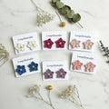 Six pairs of handmade crocheted flower stud earrings on white display cards, arranged on a light marble surface with dried flowers and greenery. The cards are labeled Loopellestudio and show white, magenta, blush pink, blue, lavender, and dusty pink flowers with yellow centers.