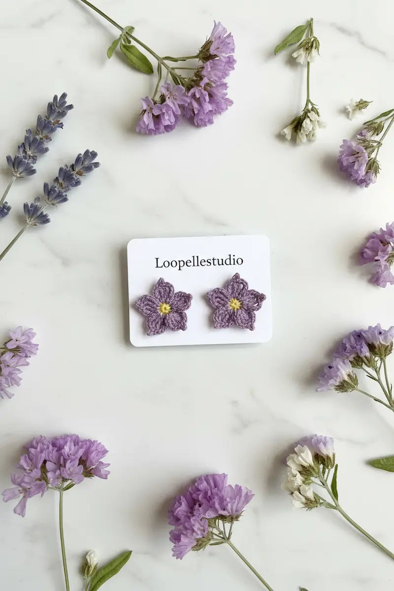 A pair of purple crocheted flower stud earrings with yellow centers, displayed on a white card labeled 'Loopellestudio' and styled on a white marble surface with purple flowers.