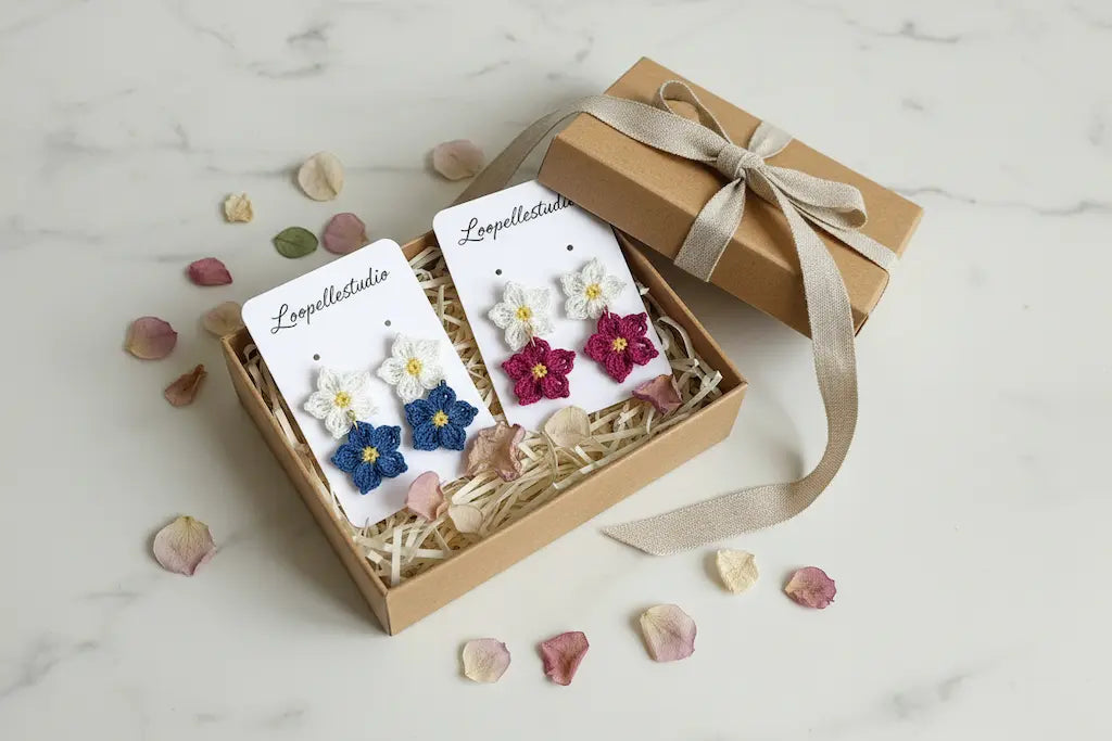 A gift box containing two pairs of handmade flower earrings displayed on white cards, with a brown kraft gift box and ribbon beside it on a light marble surface with scattered flower petals.