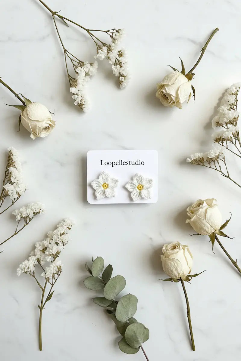A pair of white daisy stud earrings with yellow centers displayed on a small white card labeled 'Loopellestudio', arranged on a light marble surface with dried white roses, baby's breath, and eucalyptus sprigs.