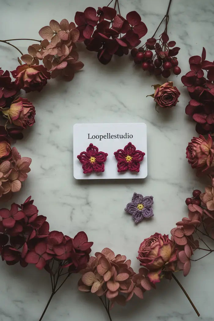 A pair of handmade crocheted magenta flower stud earrings with yellow centers on a white display card labeled 'Loopellestudio', styled on a light marble surface surrounded by a circular wreath of dried pink and burgundy flowers.