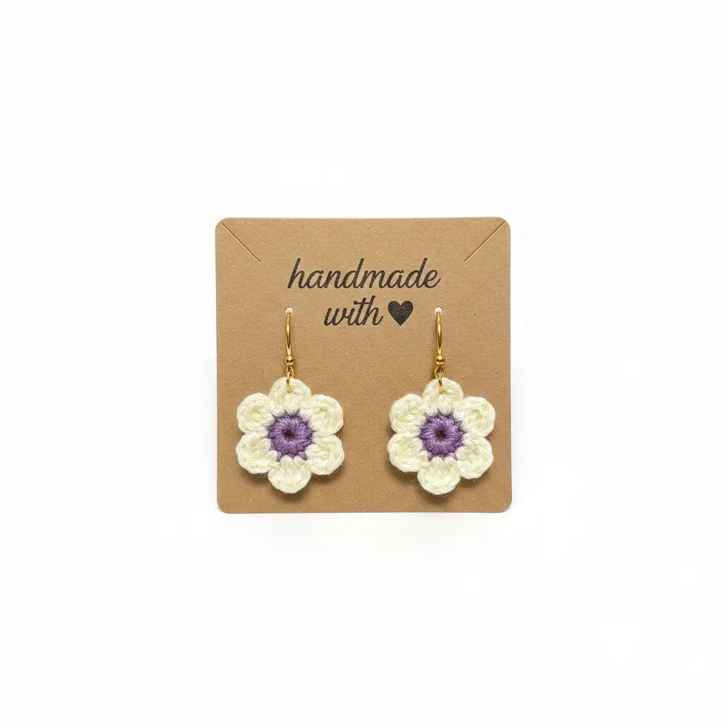 A pair of handmade crocheted flower earrings with cream petals and purple centers, hanging from gold hooks on a brown kraft display card that reads 'handmade with'.
