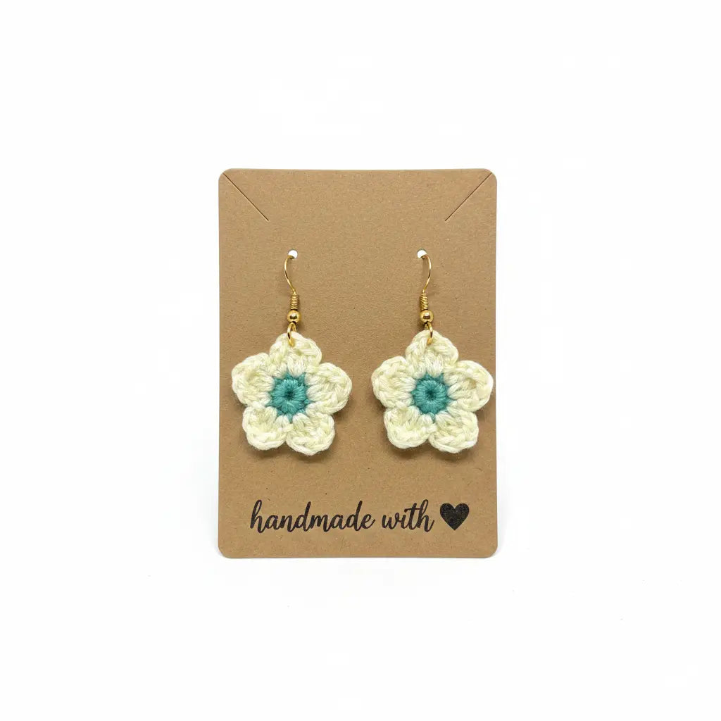 A pair of handmade crocheted flower drop earrings with cream petals and teal centers, hanging from gold hooks on a brown kraft display card that reads 'handmade with' on a white background.