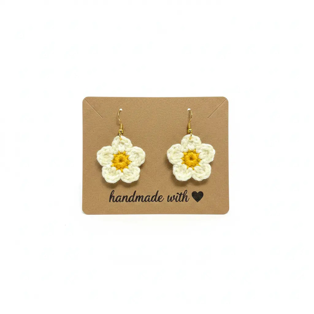 A pair of handmade crocheted daisy earrings with white petals and yellow centers, displayed on a brown kraft card that reads 'handmade with heart'.