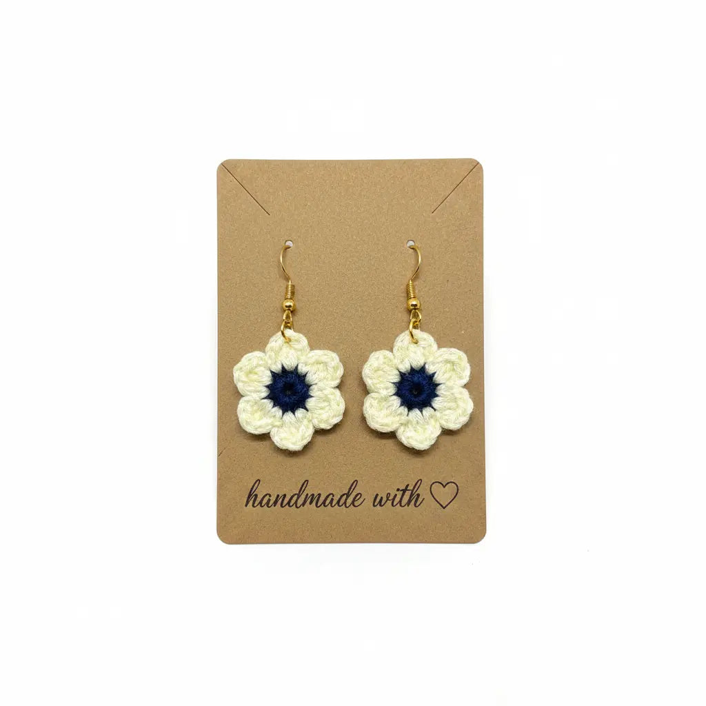 A pair of handmade crocheted dangle earrings shaped like white flowers with dark blue centers, displayed on a brown kraft card that reads 'handmade with' on a white background.
