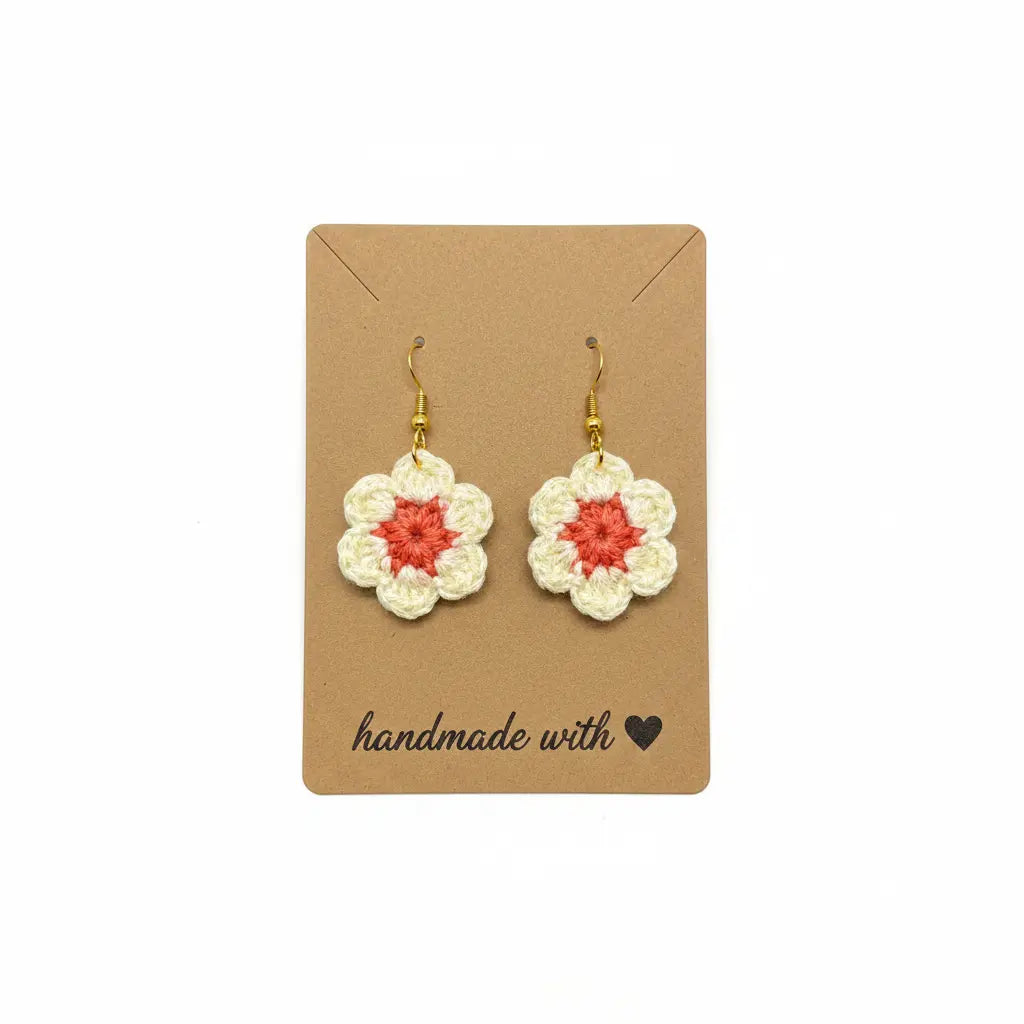 A pair of handmade crochet flower dangle earrings with cream petals, coral centers, and gold fishhook wires, displayed on a brown kraft card that reads 'handmade with' on a white background.