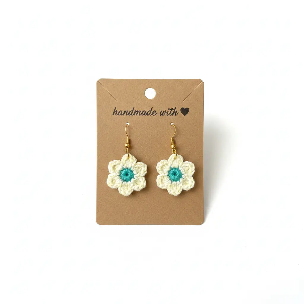 A pair of handmade crocheted flower drop earrings with cream petals, turquoise centers, and gold hooks, displayed on a brown kraft card that reads 'handmade with'.