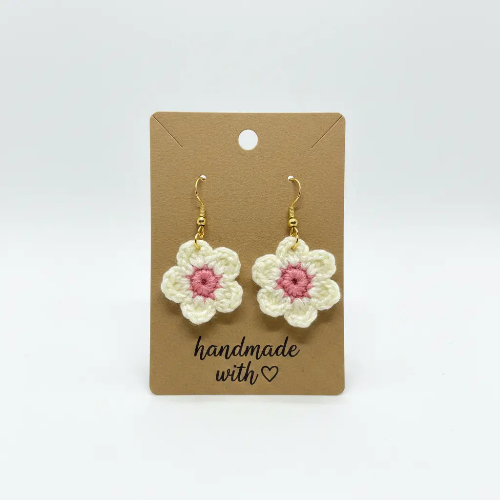 A pair of handmade crocheted flower drop earrings with white petals, pink centers, and gold hooks, displayed on a brown kraft card that reads 'handmade with heart'.
