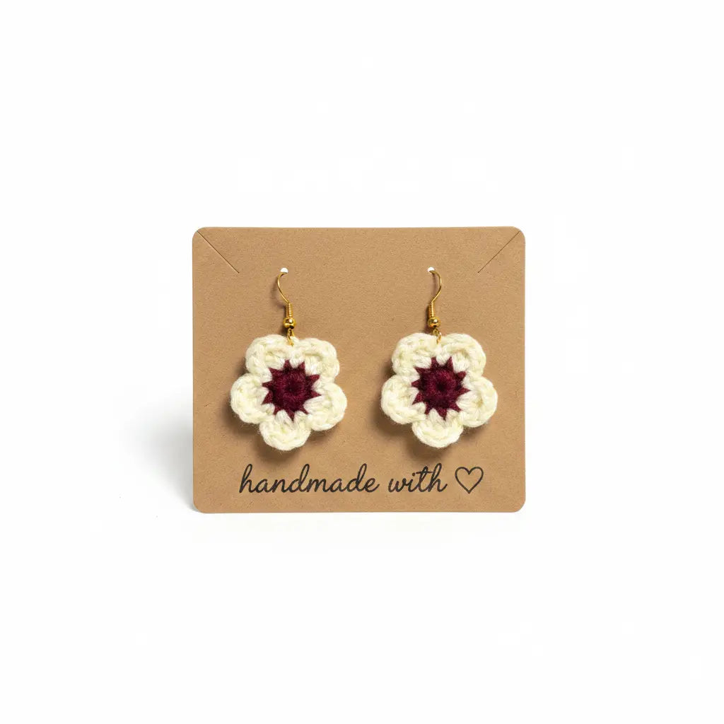 A pair of handmade crocheted flower drop earrings with cream petals and dark red centers, displayed on a brown kraft card that reads 'handmade with heart'.