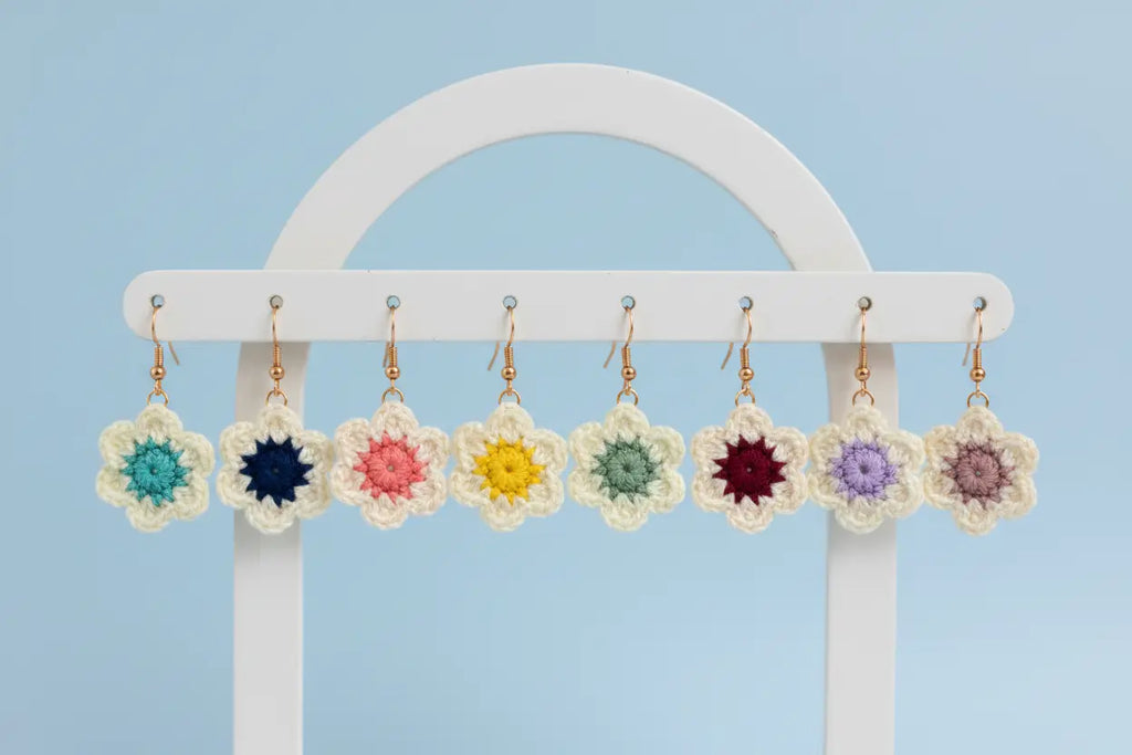 A set of handmade crochet flower earrings in various colors, hanging from a white metal display stand against a light blue background.