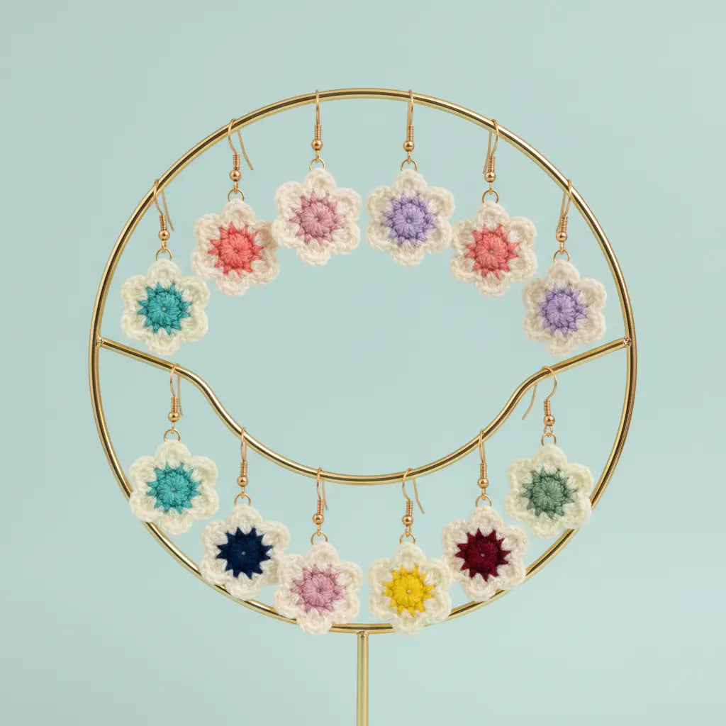 A set of crocheted flower-shaped earrings displayed on a gold circular stand against a light blue background. The earrings feature cream-colored outer petals and colorful centers in pink, purple, teal, navy, yellow, and burgundy.