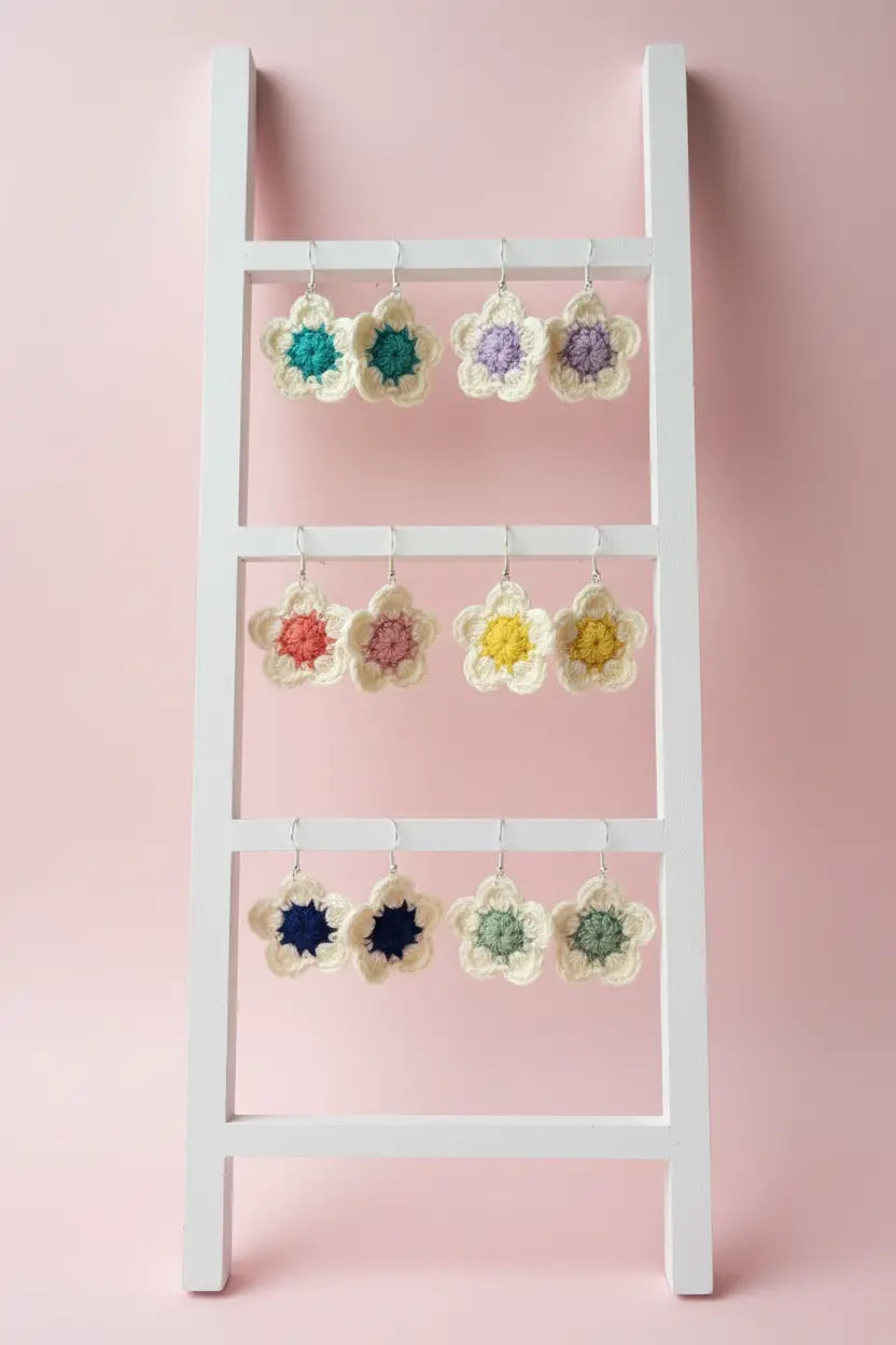 A white wooden ladder-style hanger with three horizontal bars displays multiple crocheted flower-shaped earrings in assorted colors against a soft pink background.