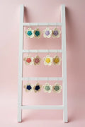 A white wooden ladder-style hanger with three horizontal bars displays multiple crocheted flower-shaped earrings in assorted colors against a soft pink background.
