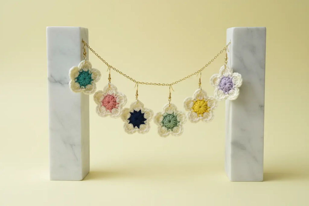 A gold chain necklace with six crocheted flower charms in pastel colors, displayed on a marble stand against a pale yellow background.