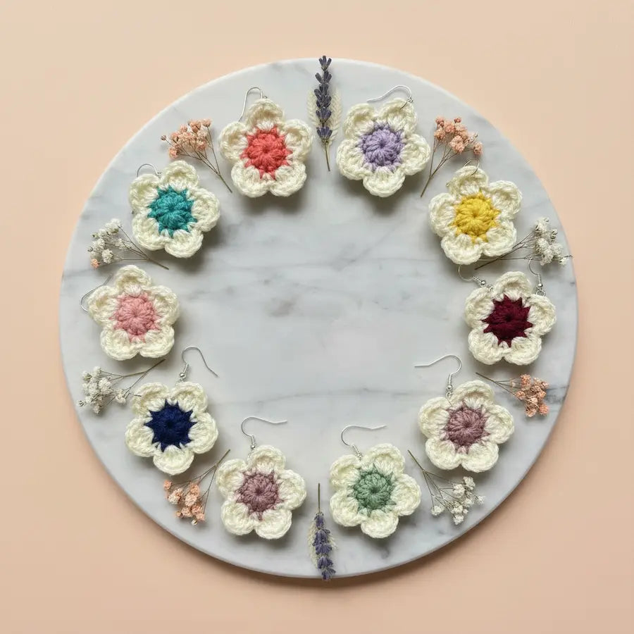 A circular arrangement of handmade crocheted flower earrings in multiple colors, displayed on a round white marble tray with dried flowers against a soft peach background.