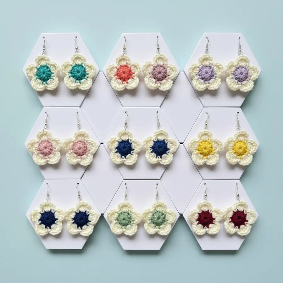 Twelve pairs of handmade crochet flower earrings displayed on white hexagonal cards against a light blue background. The earrings feature cream-colored petals with various colorful centers in teal, coral, lavender, pink, navy, yellow, green, and red.