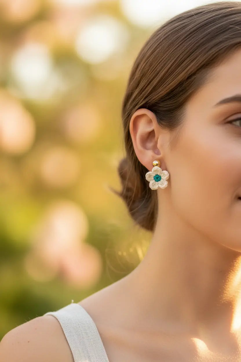 A close-up lifestyle shot of a woman wearing a delicate gold flower-shaped stud earring with a green center and white petals, set against a warm, blurred bokeh background.