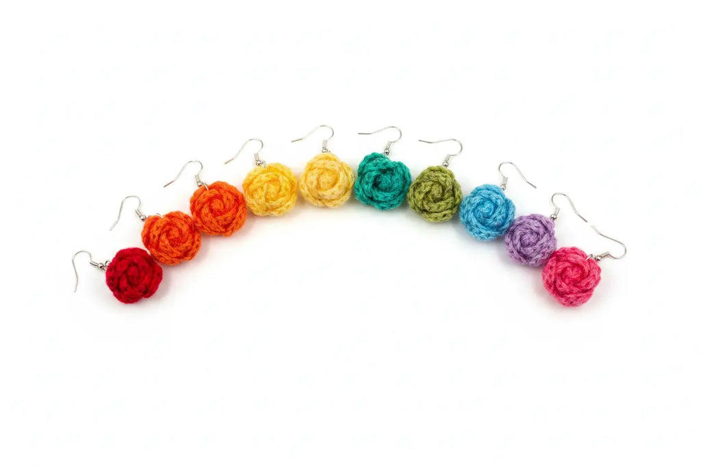 A set of handmade crochet flower drop earrings arranged in a rainbow sequence on a white background. The earrings feature silver-toned French hook backs and are made of textured yarn in red, orange, yellow, green, blue, purple, and pink.
