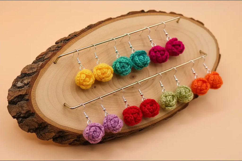 A set of colorful crochet flower dangle earrings displayed on a rustic wood slice stand against a solid peach background.