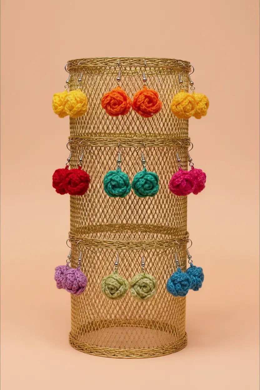 A gold wire mesh cylindrical display stand holding multiple pairs of colorful crocheted flower-shaped earrings in yellow, orange, red, teal, pink, purple, green, and blue, set against a solid peach background.