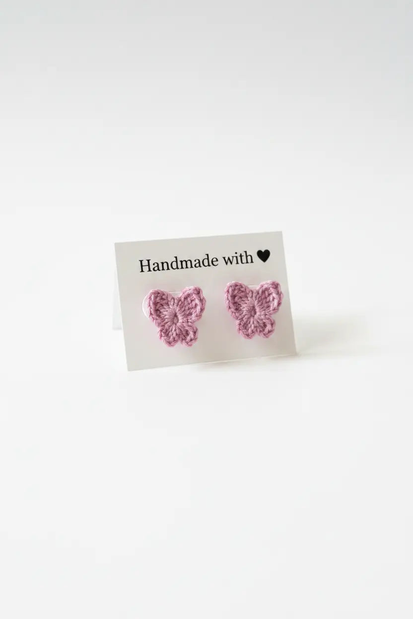 A pair of pink crocheted butterfly stud earrings displayed on a small white card that reads 'Handmade with' against a plain white background.
