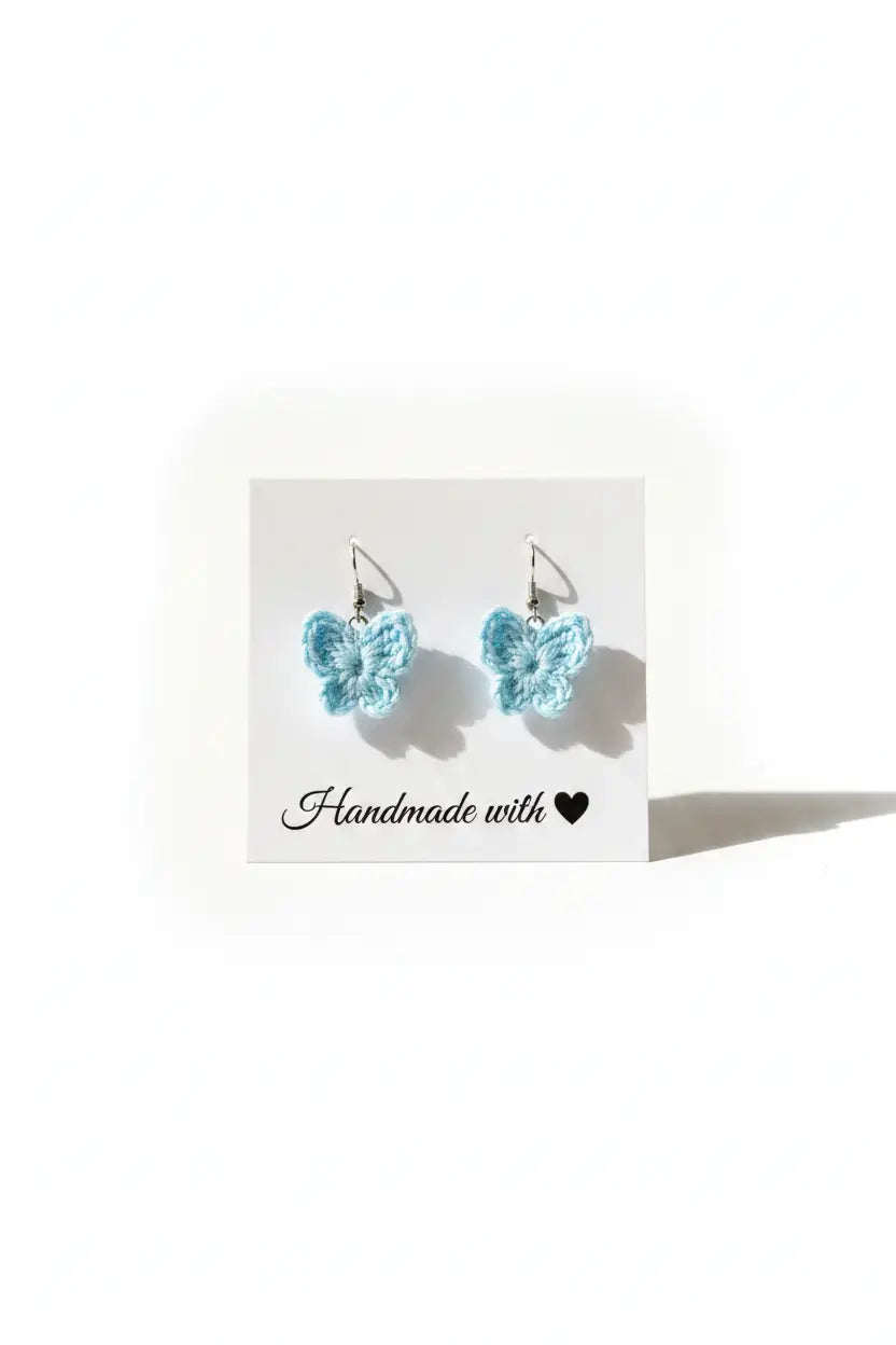 A pair of light blue crocheted butterfly earrings on a white display card that reads 'Handmade with a heart', set against a plain white background.
