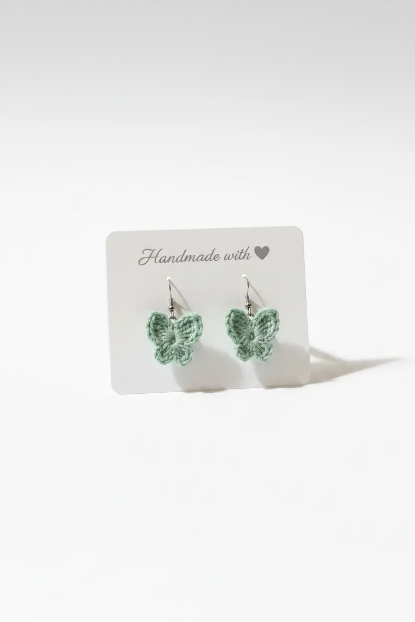 A pair of small crocheted butterfly earrings in a light mint green color, displayed on a white card that reads 'Handmade with' against a clean white background.
