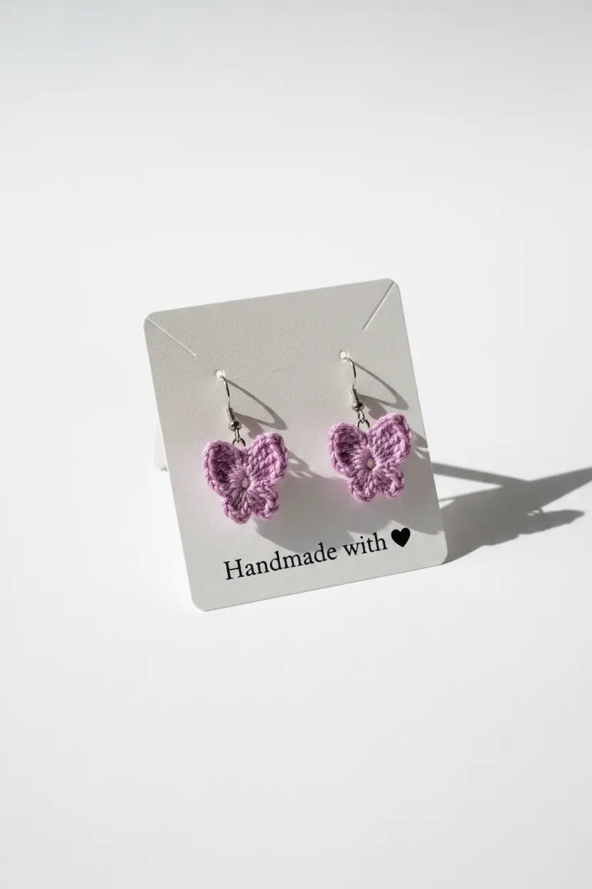 A pair of small crocheted butterfly earrings in light purple pink, displayed on a white card that reads 'Handmade with' on a clean white background.