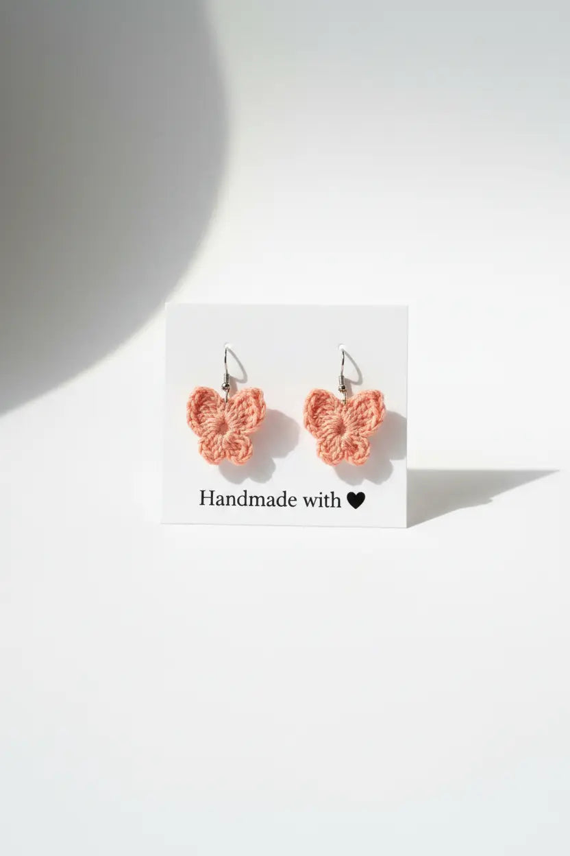 A pair of peach-pink crocheted butterfly earrings on a white display card that reads 'Handmade with a heart', set against a bright white background with soft shadows.