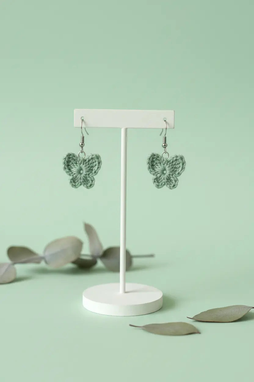 A pair of green crocheted butterfly earrings displayed on a white stand against a mint green background with scattered eucalyptus leaves.