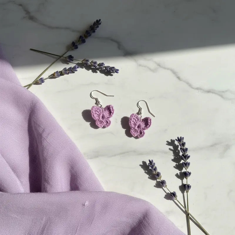 A pair of small lavender crocheted butterfly earrings with silver fishhook wires, displayed on a white marble surface with decorative lavender sprigs and a light purple fabric drape.