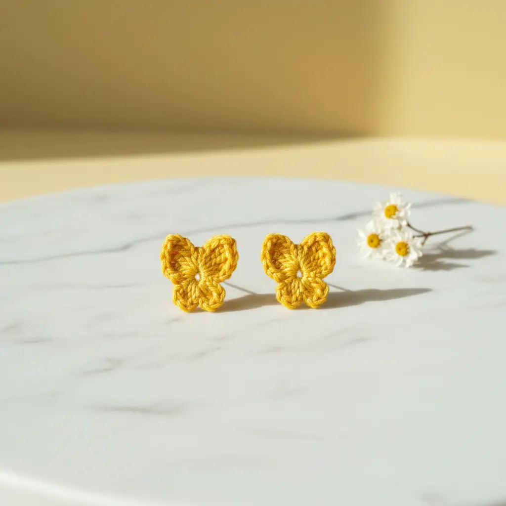A pair of yellow crocheted butterfly stud earrings displayed on a white marble surface, with a small white daisy prop in the background against a warm yellow wall.