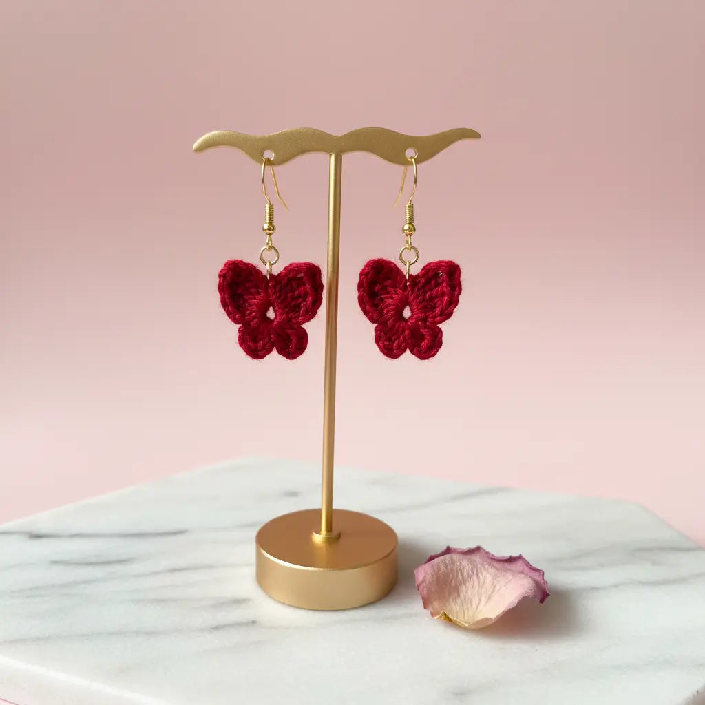 A pair of red crocheted butterfly drop earrings displayed on a gold stand against a soft pink background, with a marble surface and a dried rose petal nearby.