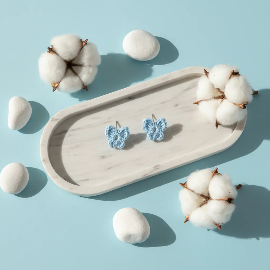 A pair of small light blue crocheted butterfly-shaped earrings displayed on a light wood tray, surrounded by cotton bolls and white stones on a light blue background.