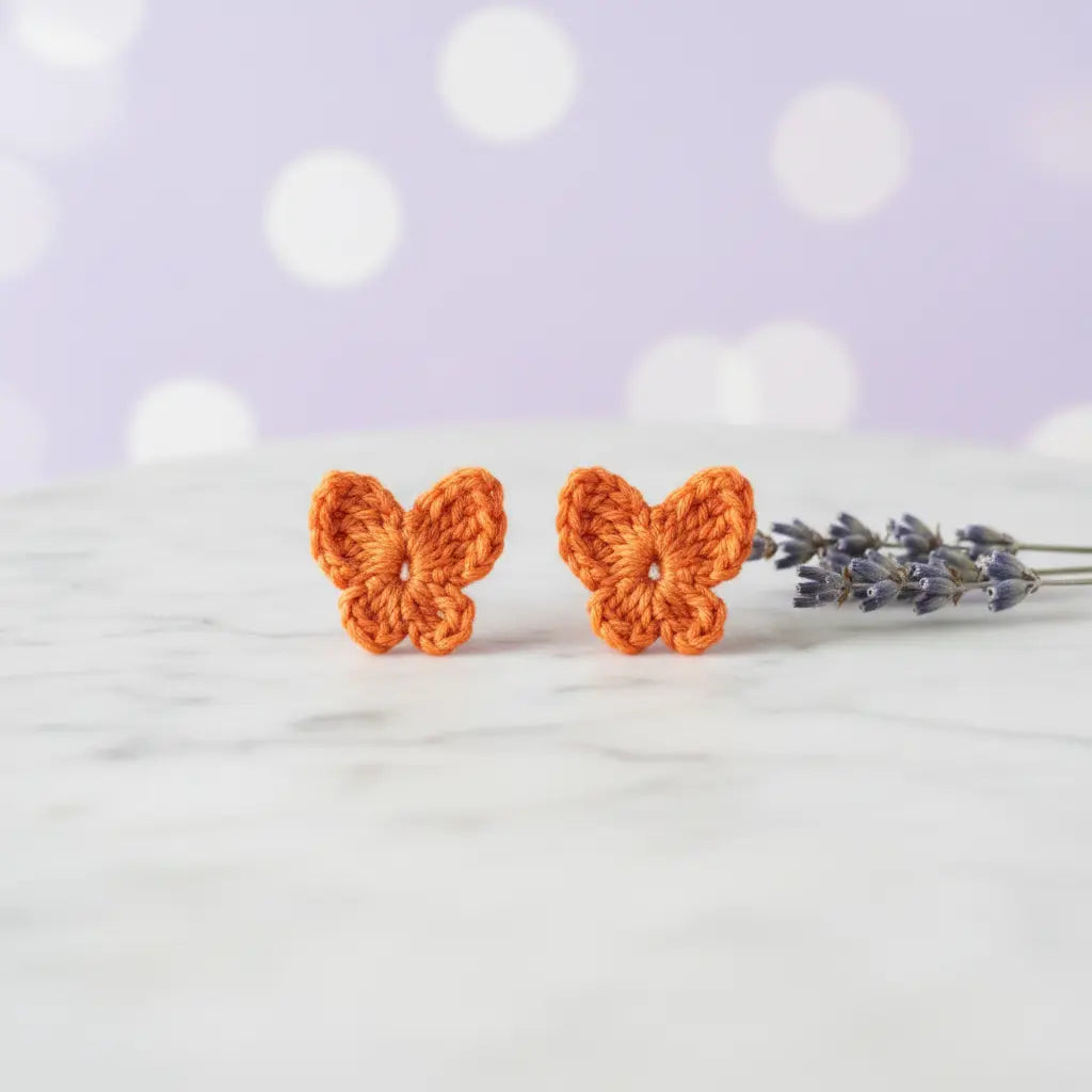 A pair of small crocheted butterfly stud earrings in bright orange, displayed on a white marble surface next to a sprig of lavender against a soft purple bokeh background.