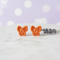 A pair of small crocheted butterfly stud earrings in bright orange, displayed on a white marble surface next to a sprig of lavender against a soft purple bokeh background.