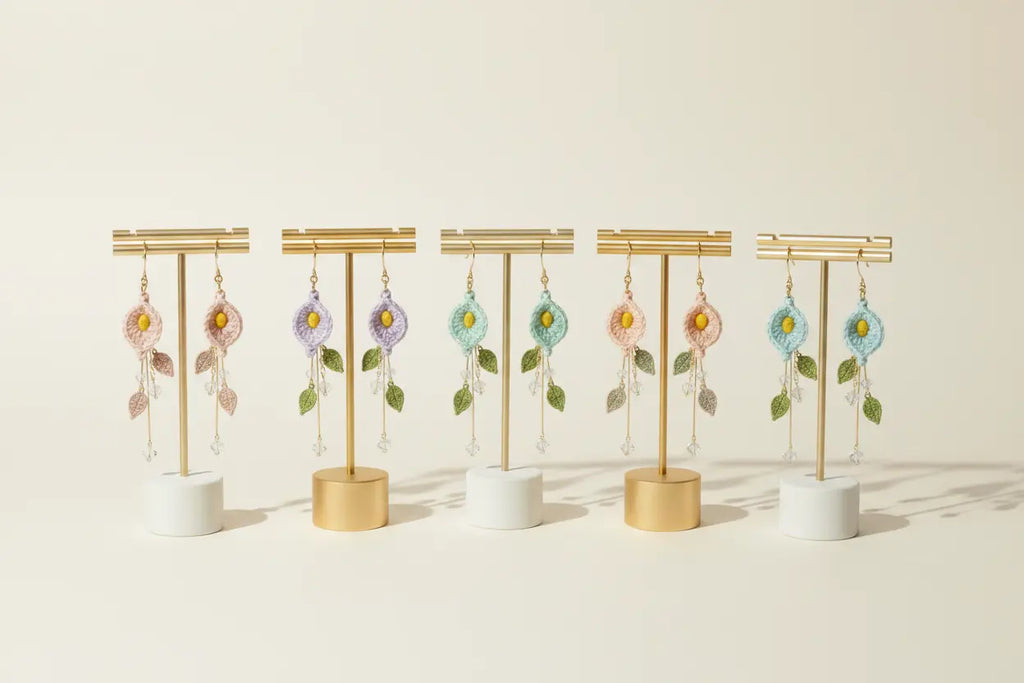 Six pairs of handmade beaded dangle earrings displayed on gold T-stands with white and gold cylindrical bases, arranged in a row against a warm cream background.