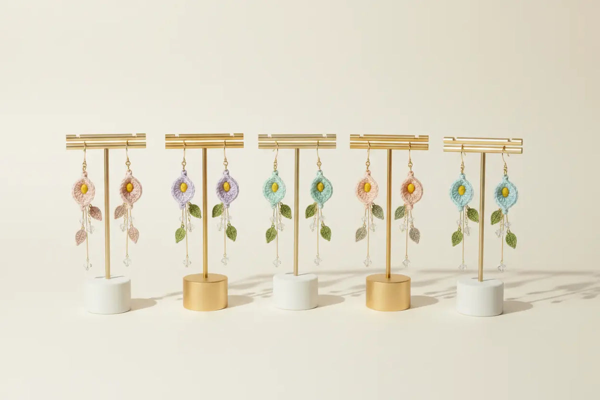Six pairs of handmade beaded dangle earrings displayed on gold T-stands with white and gold cylindrical bases, arranged in a row against a warm cream background.