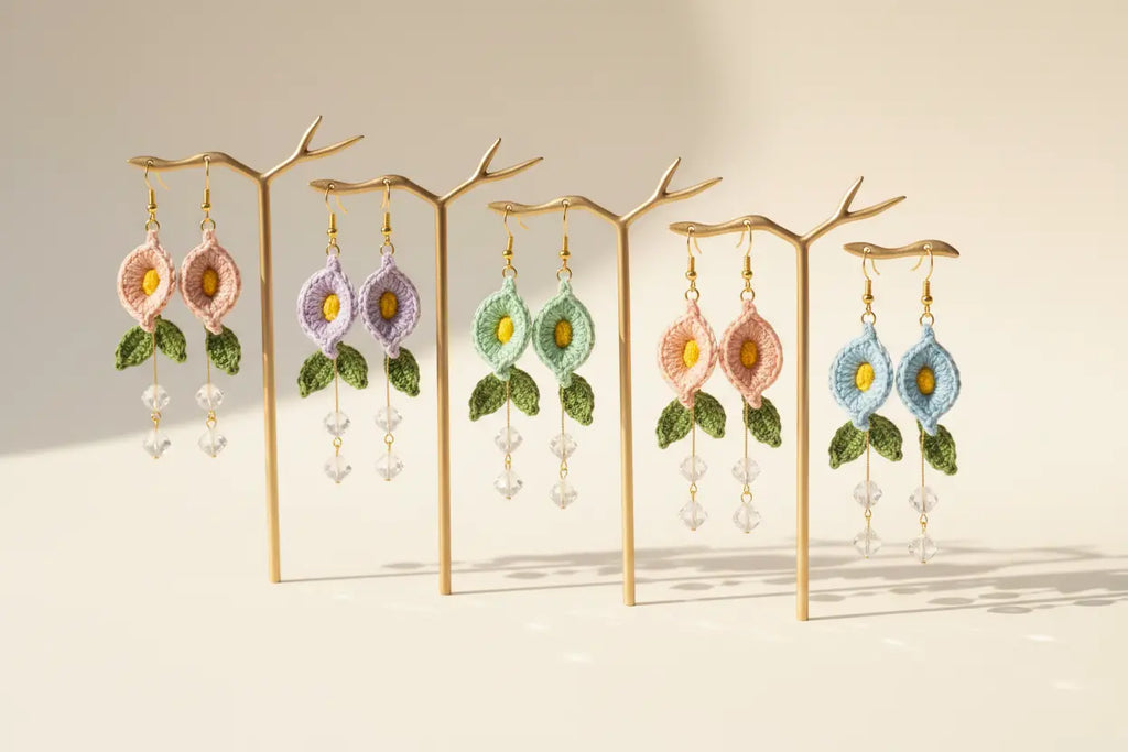 Six pairs of handmade floral dangle earrings displayed on gold branch-shaped stands against a warm beige background. The earrings feature embroidered flowers in pink, purple, mint green, peach, and light blue, with green leaves and dangling clear crystal beads.